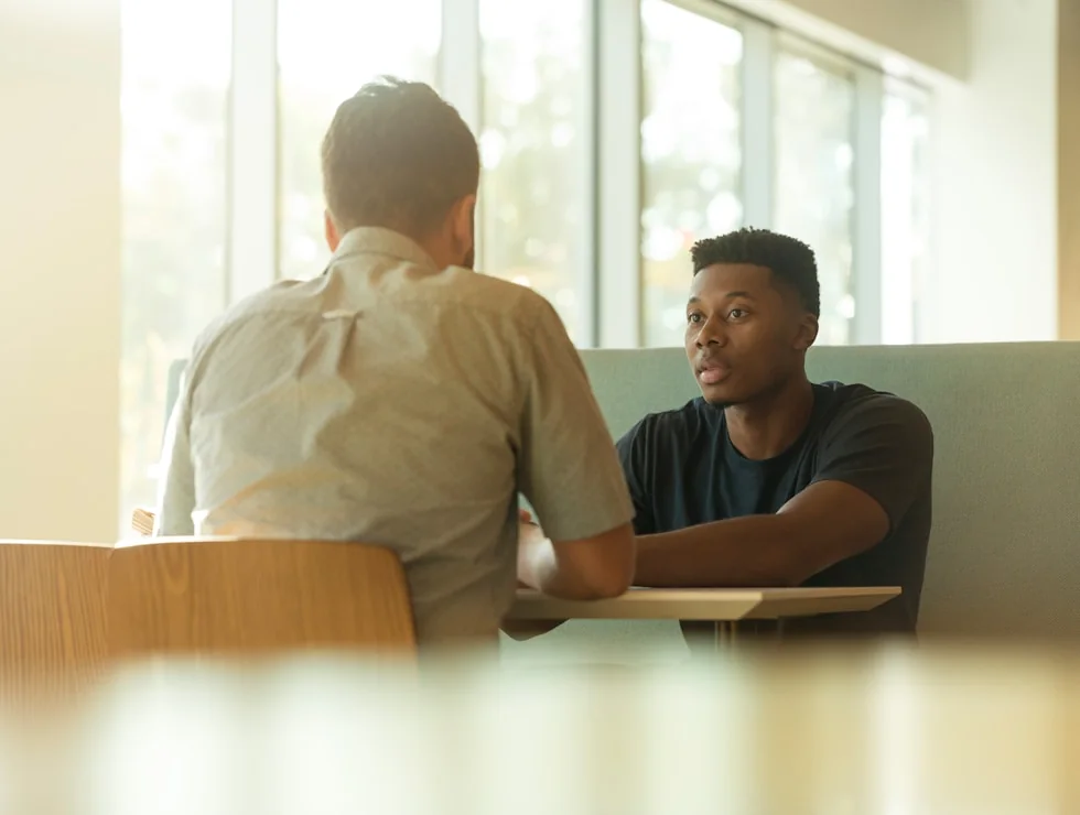 Two professionals in conversation across a table by a window.